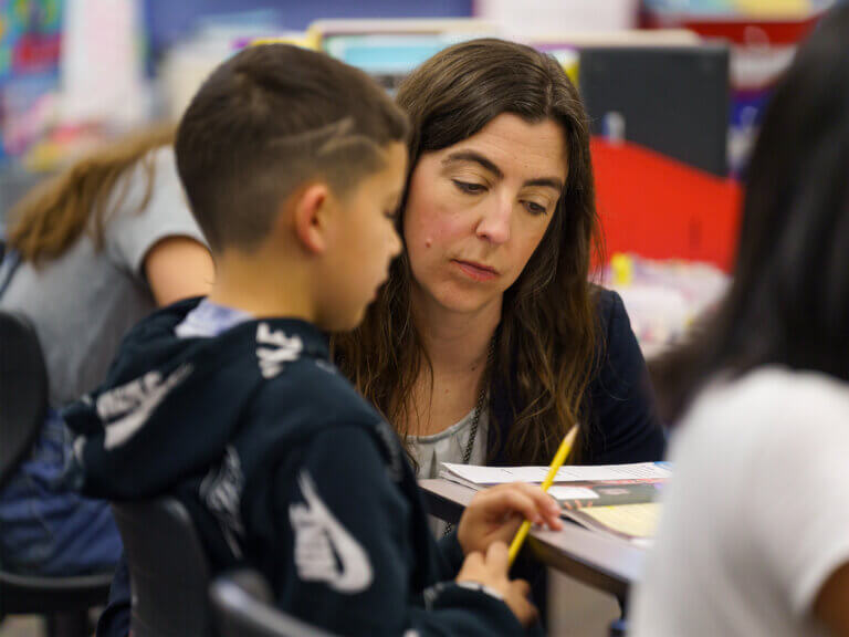 A teacher crouches next to a student at the student's desk.